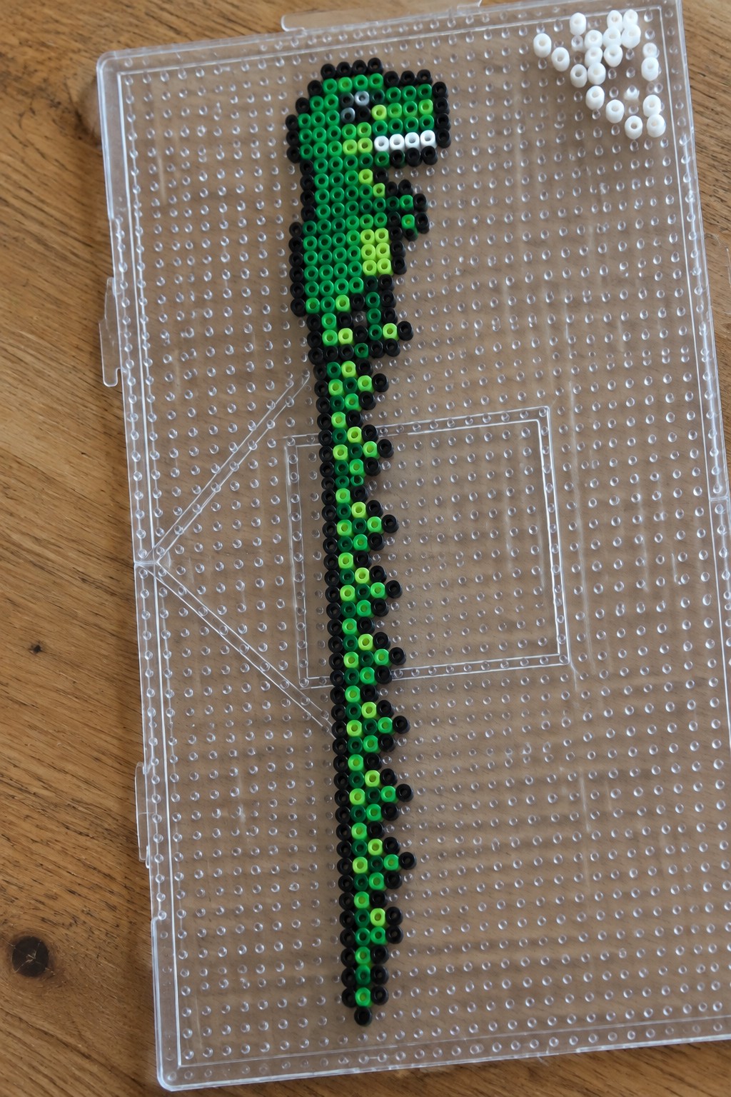 Green and black perler bead snake on clear pegboard with loose white beads nearby
