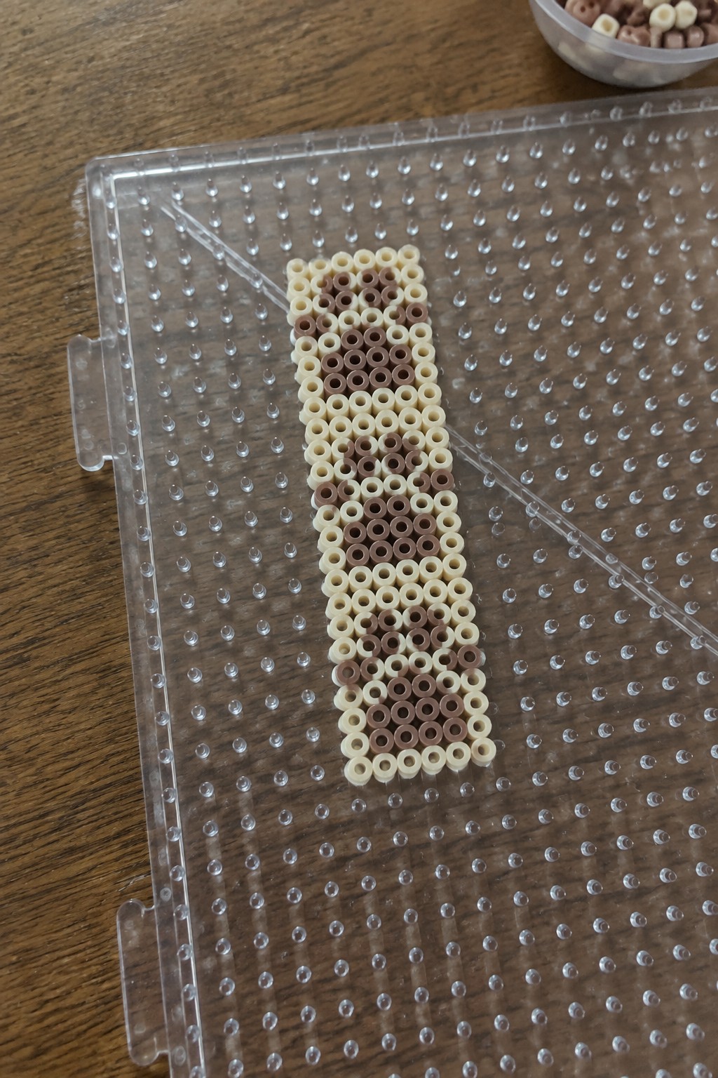 Brown and cream perler beads patterned vertically on clear pegboard over wooden table