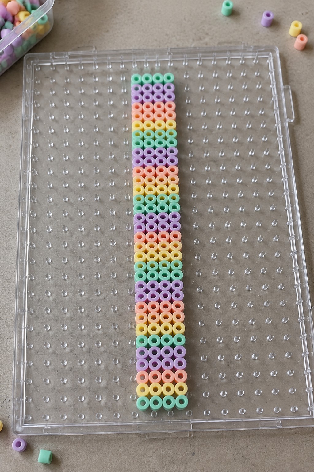 Clear pegboard with vertical pastel beads in rainbow stripes and scattered loose beads.