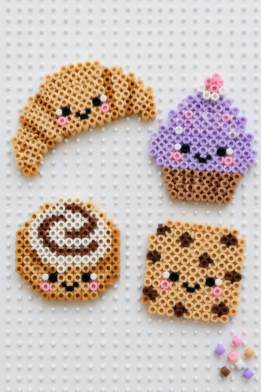Four unfused Perler bead kawaii food characters on a pegboard showing a croissant, cupcake, round cookie, and square cookie, each with simple faces.