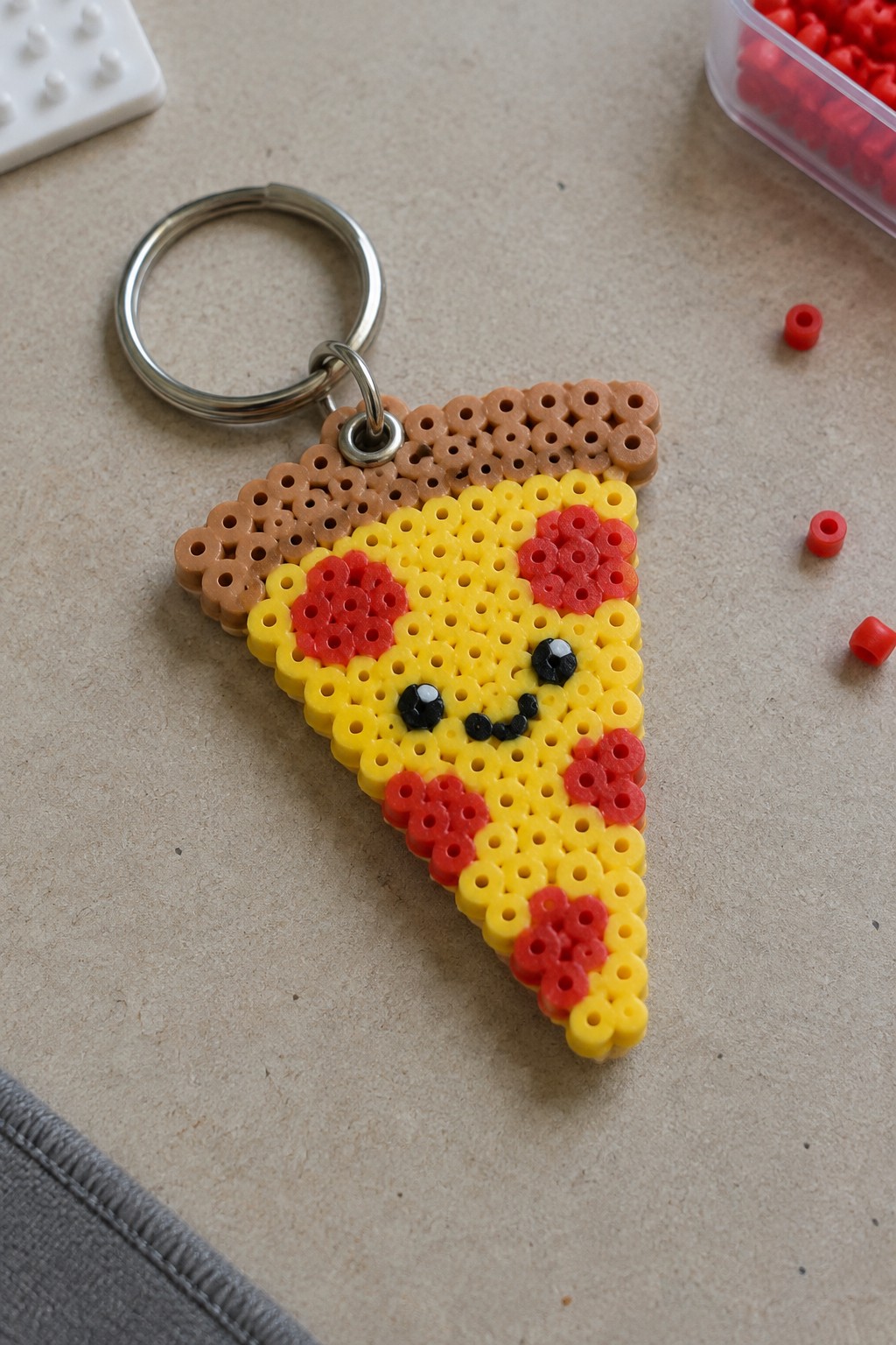 A fused Perler bead pizza slice keychain with yellow cheese, red pepperoni clusters, brown crust, and a kawaii face attached to a metal ring.