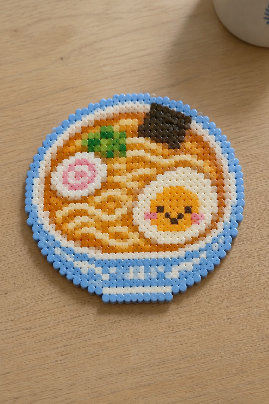 Perler bead art of cute ramen bowl with faced egg on wood table.