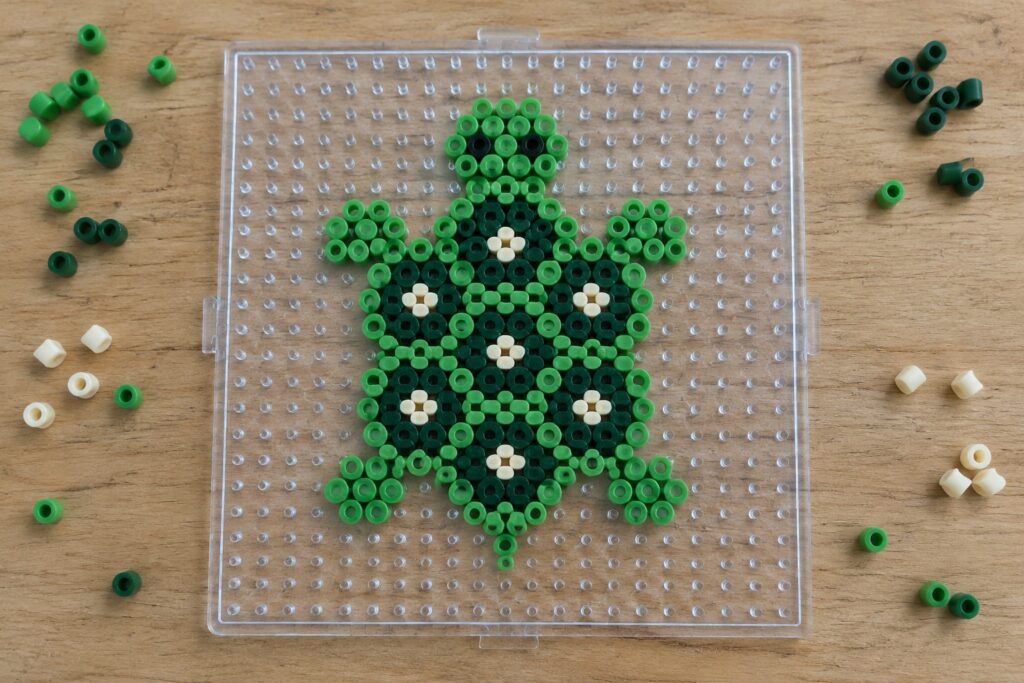 Green perler bead turtle with white flower shell on clear pegboard with loose beads