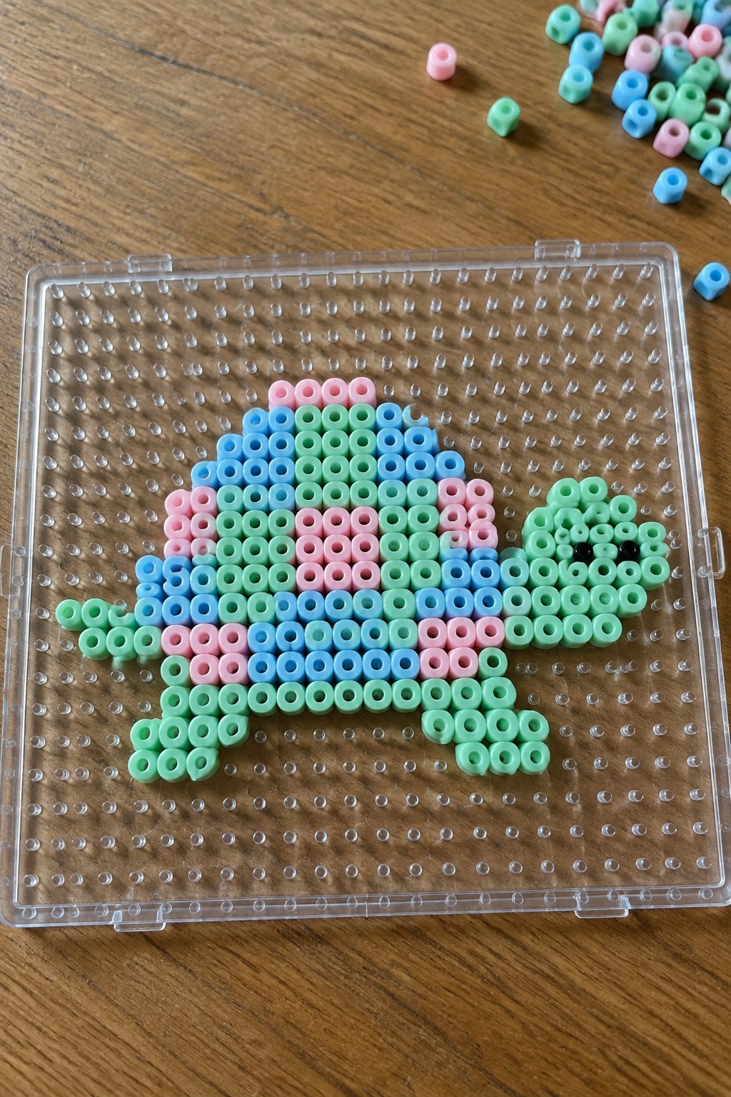 Perler bead turtle with pink blue and mint green shell on a clear pegboard.