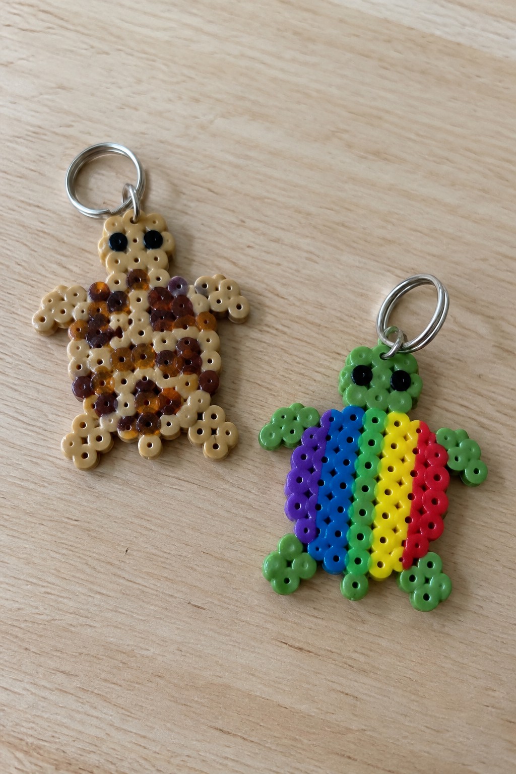 Two perler bead turtle keychains on wood, one brown mottled and one rainbow.