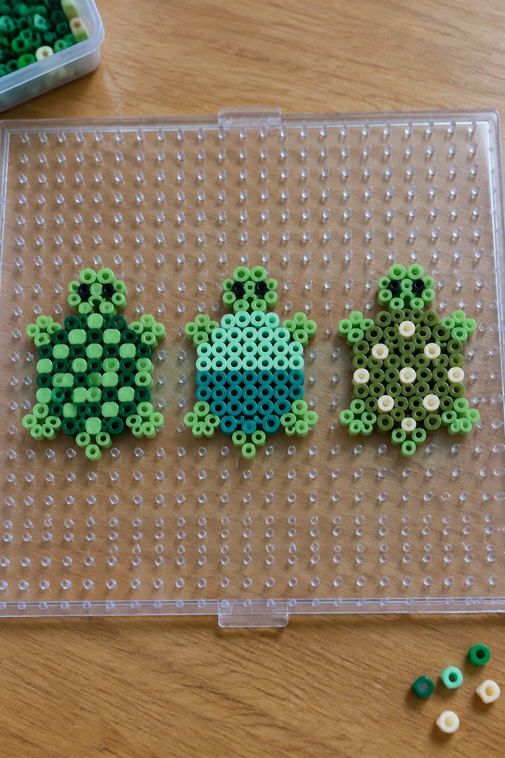 Three green perler bead frogs on a clear pegboard over wood.