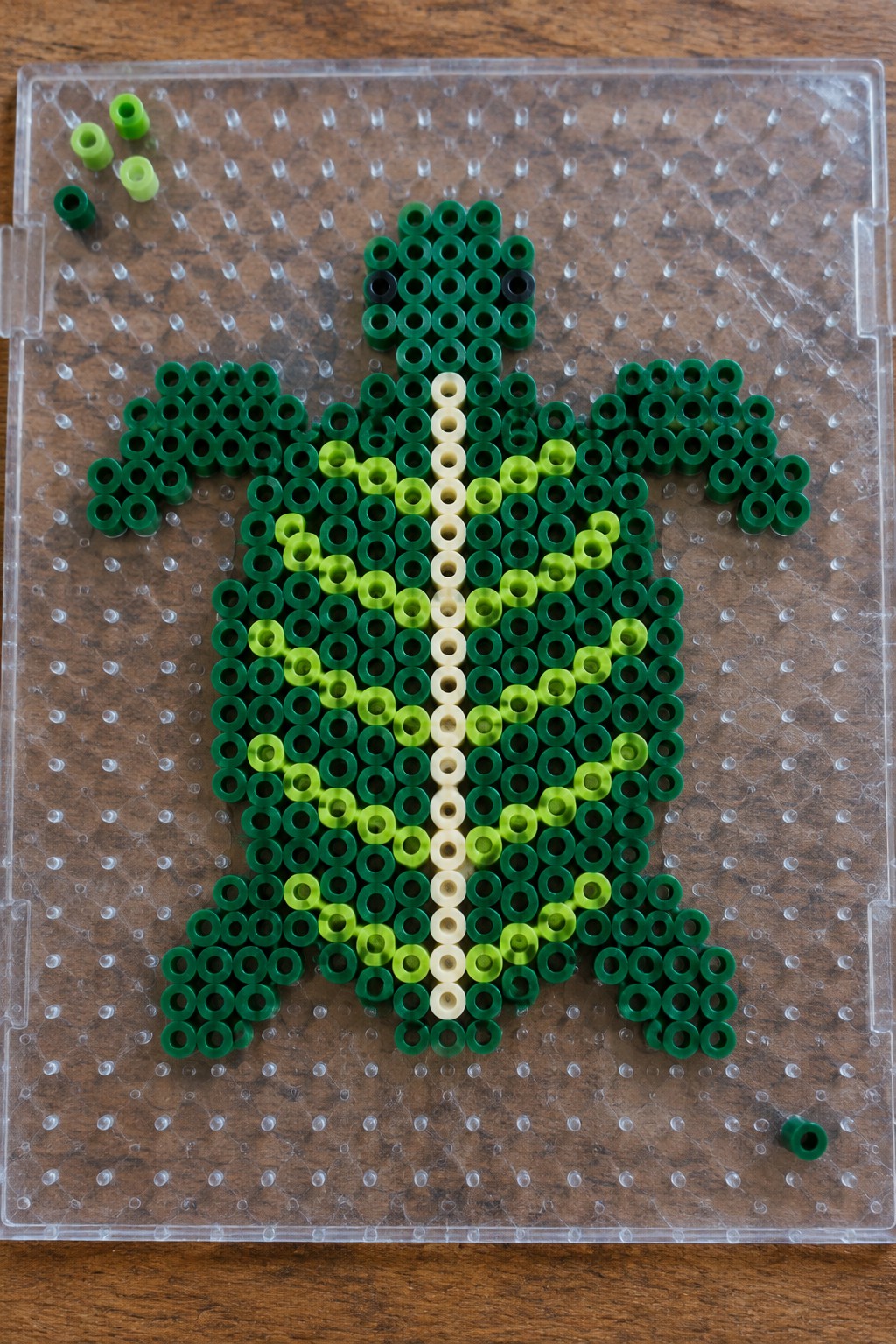 Green perler bead turtle on clear pegboard with scattered loose beads nearby