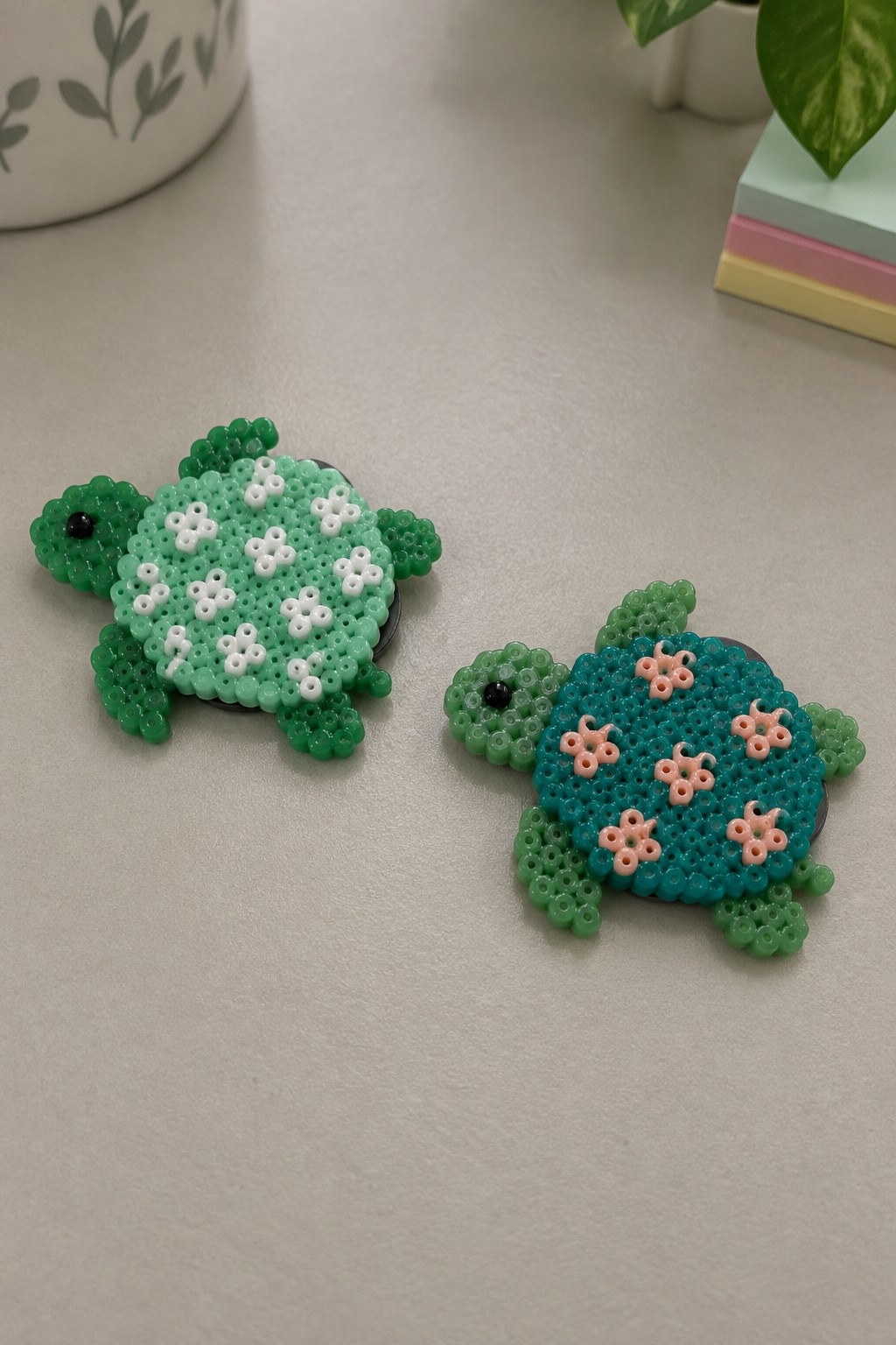 Two perler bead turtles, green with white shell and teal with pink flowers.
