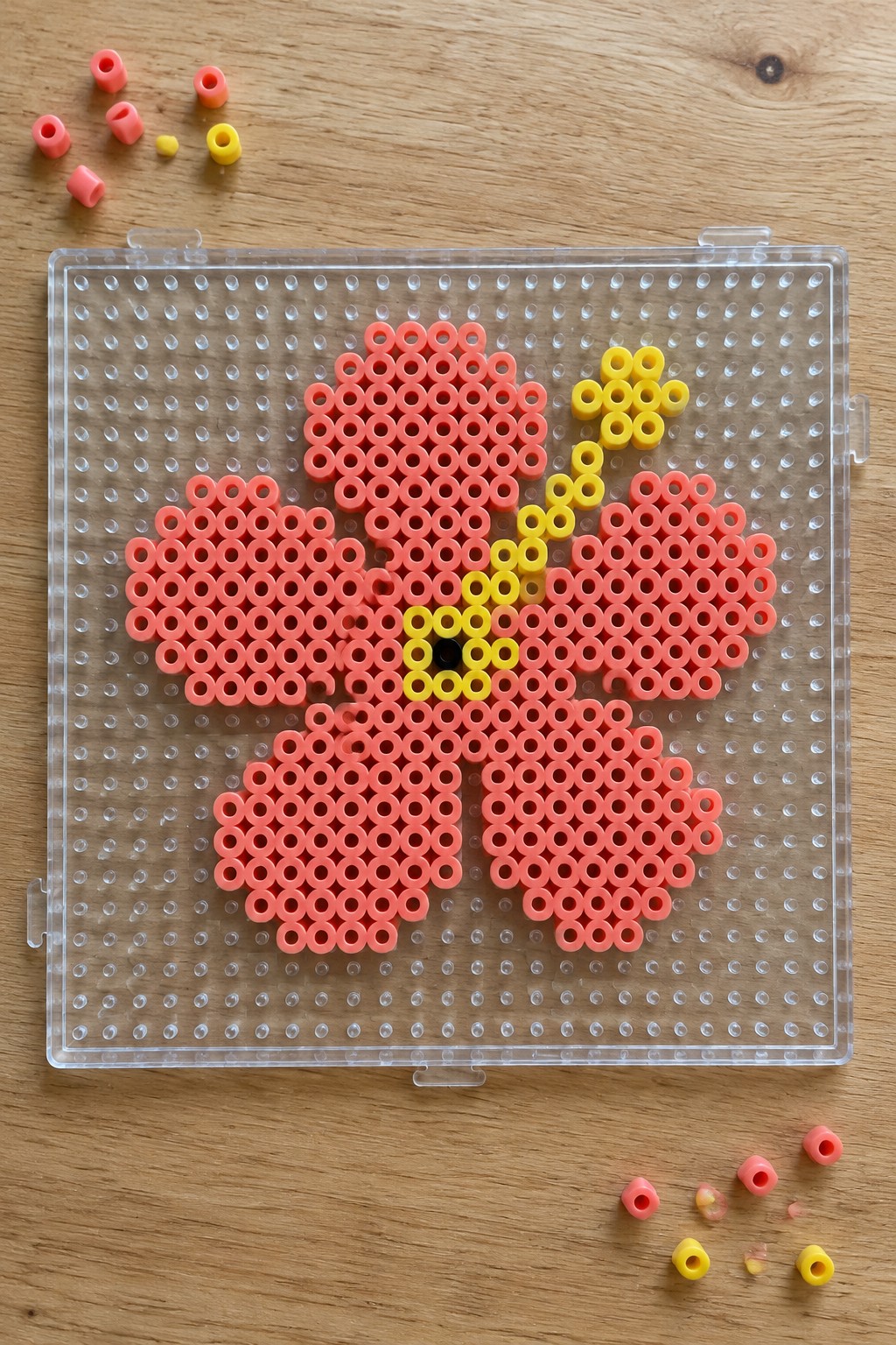 Pink and yellow perler bead flower with black center on clear pegboard.
