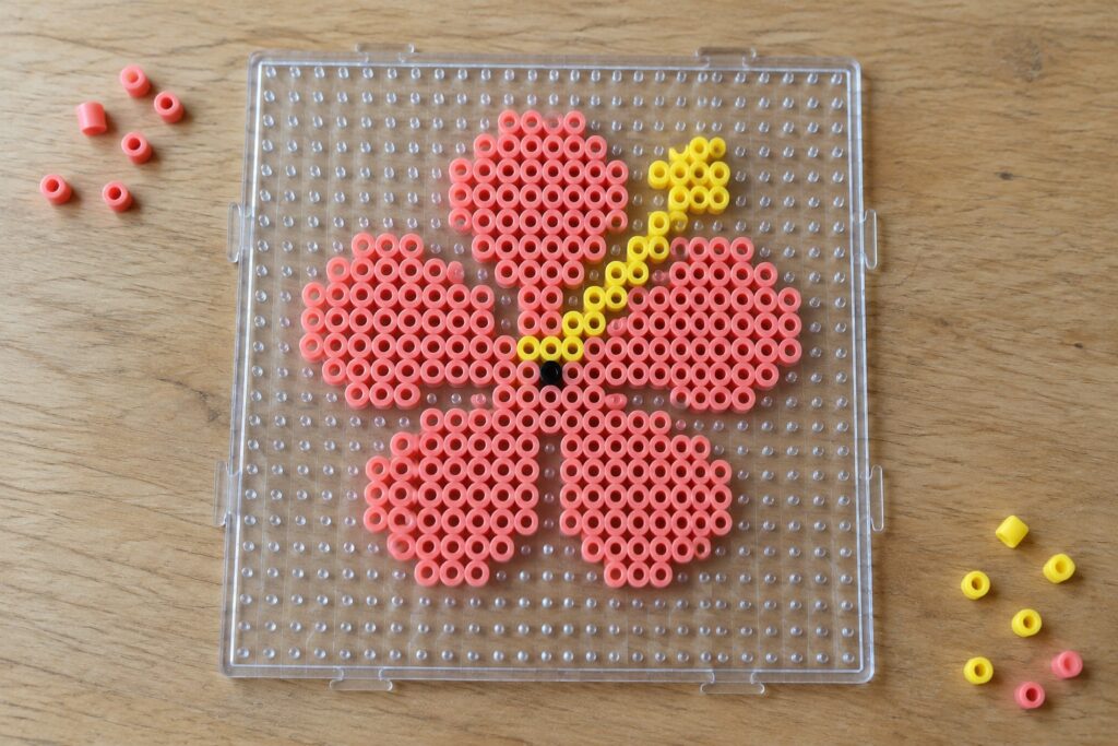 Pink and yellow perler bead flower with black center on clear pegboard.