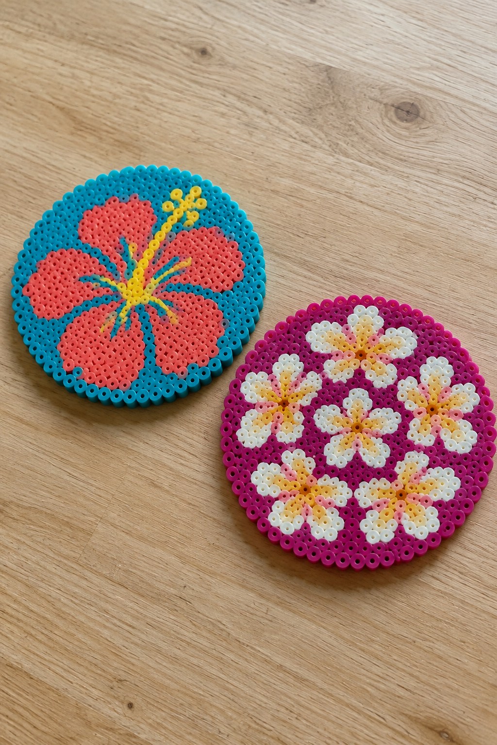 Two round Perler bead designs on wood showing a pink hibiscus on turquoise and a symmetrical magenta and yellow floral pattern.