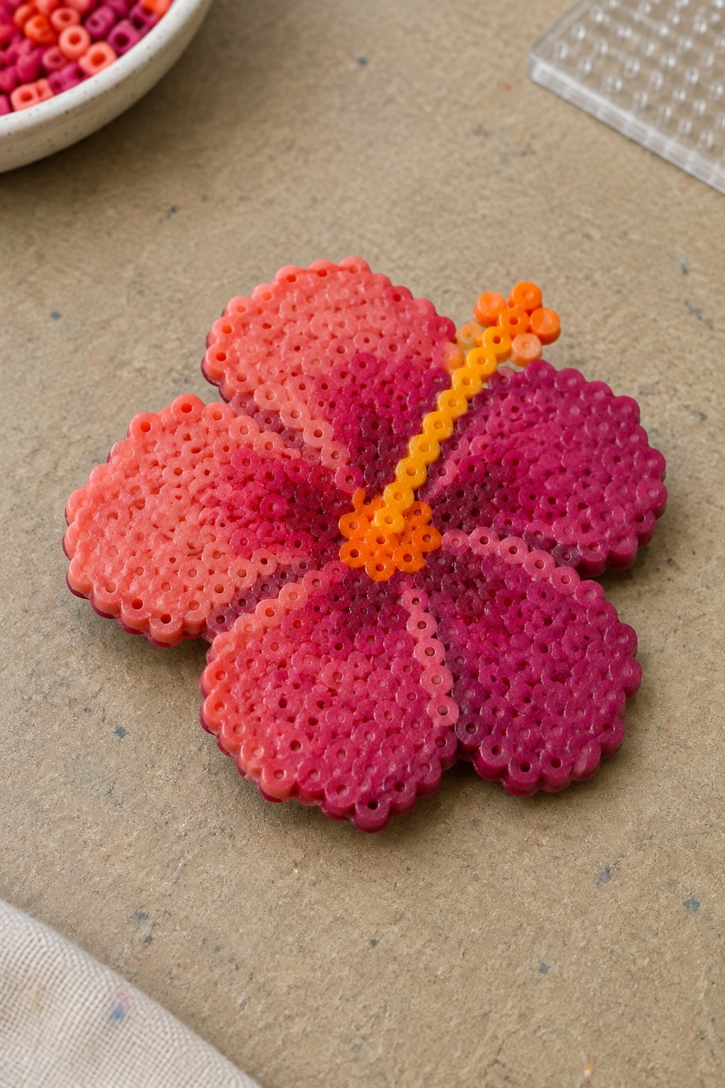 Pink and magenta perler bead hibiscus flower with orange center on table.