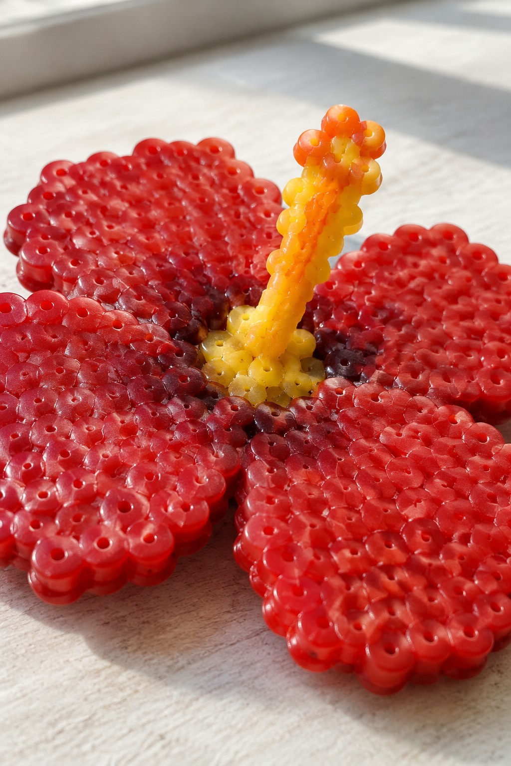 Red perler bead hibiscus flower with yellow-orange beaded center on wood