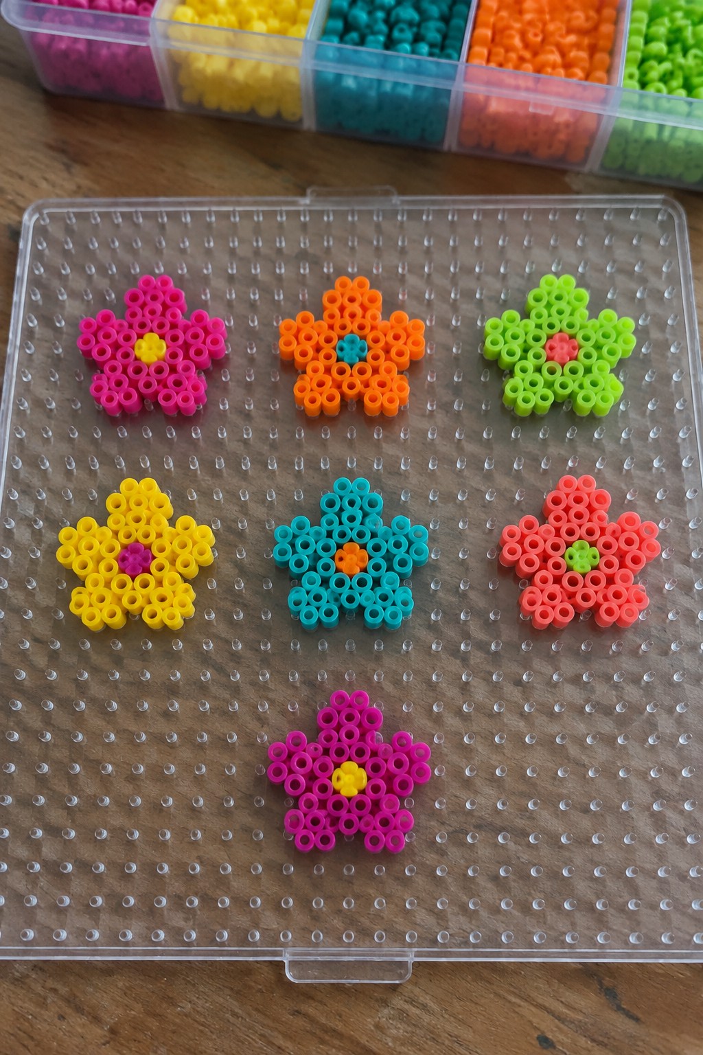 Colorful perler bead flowers arranged on clear pegboard with bead organizer above.