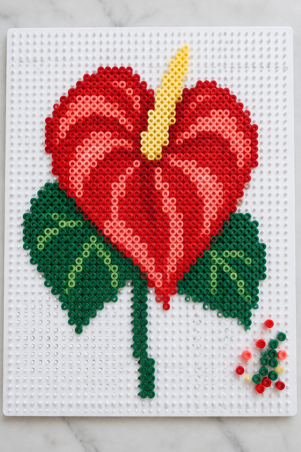 Red perler bead flower with yellow stamen and green leaves on white pegboard.