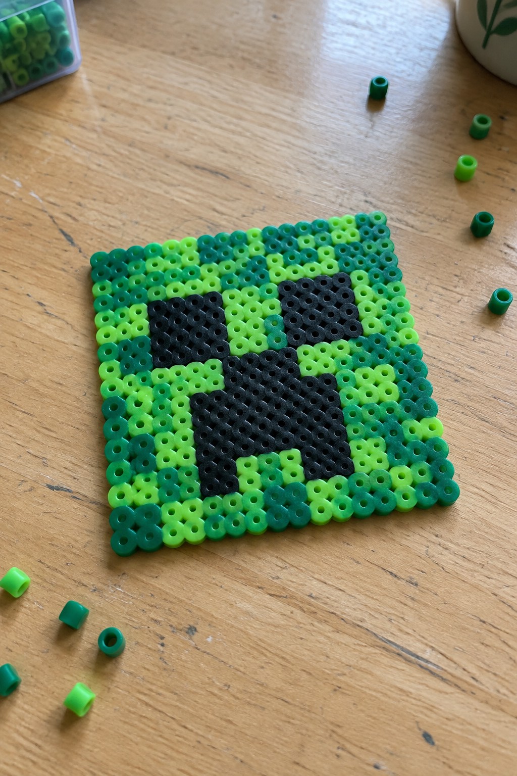 Green and black perler bead pixel art square resembling creeper on wooden table
