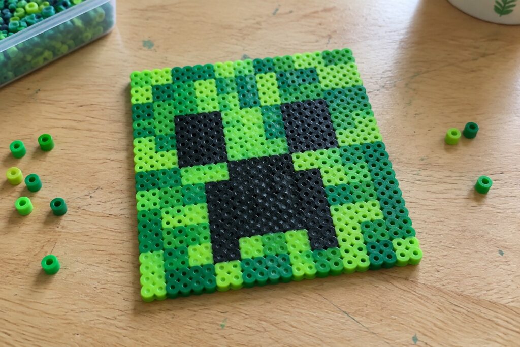 Green and black perler bead pixel art square resembling creeper on wooden table