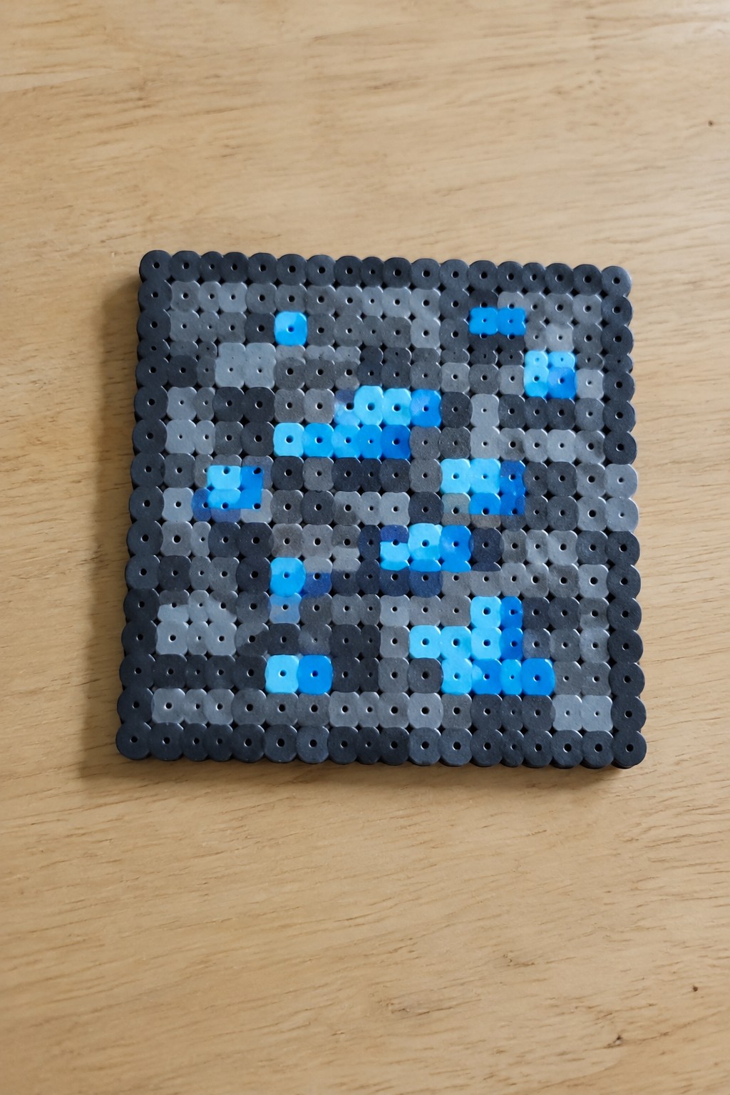 Square perler bead mosaic in shades of gray with blue pixel accents on wood