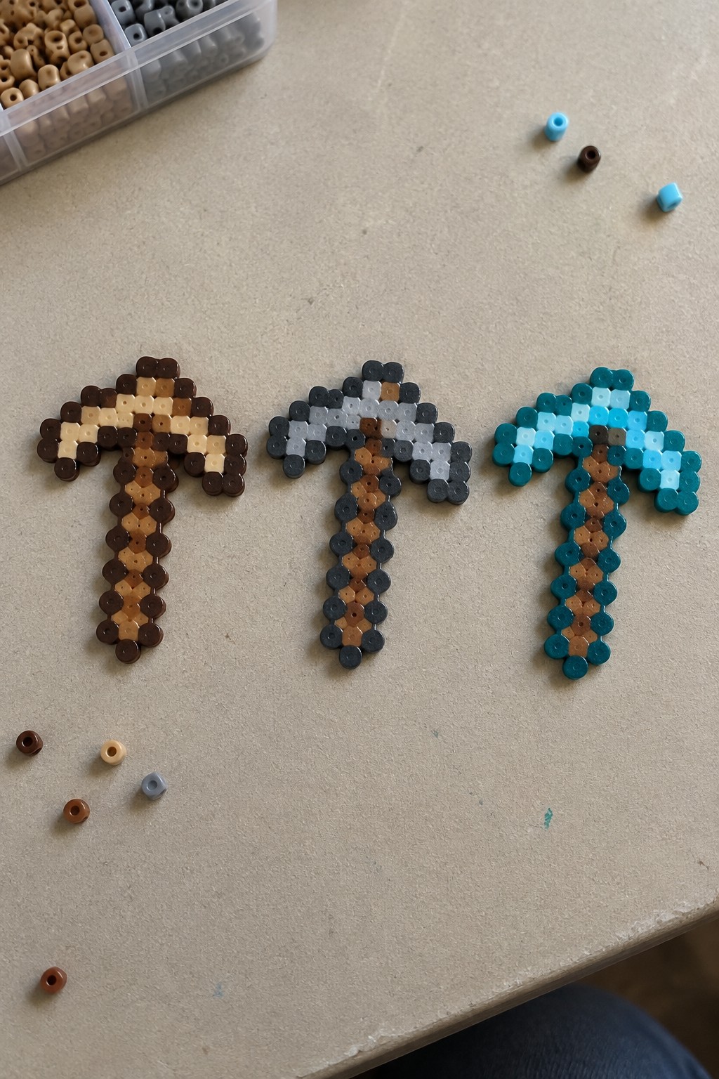 Three Perler bead Minecraft pickaxes in brown, gray, and teal with loose beads nearby