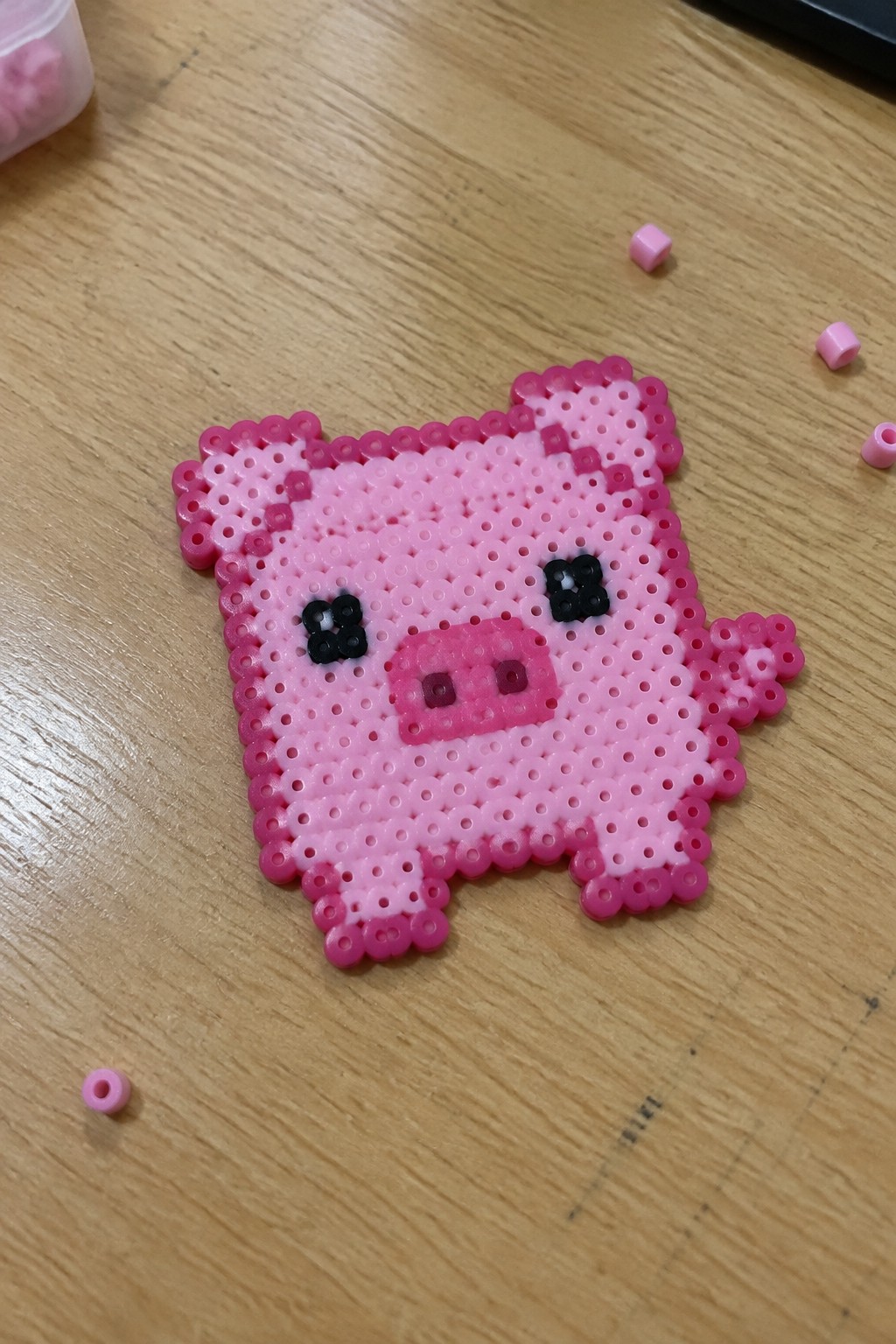 Pink perler bead pig with black eyes on wooden table with loose beads