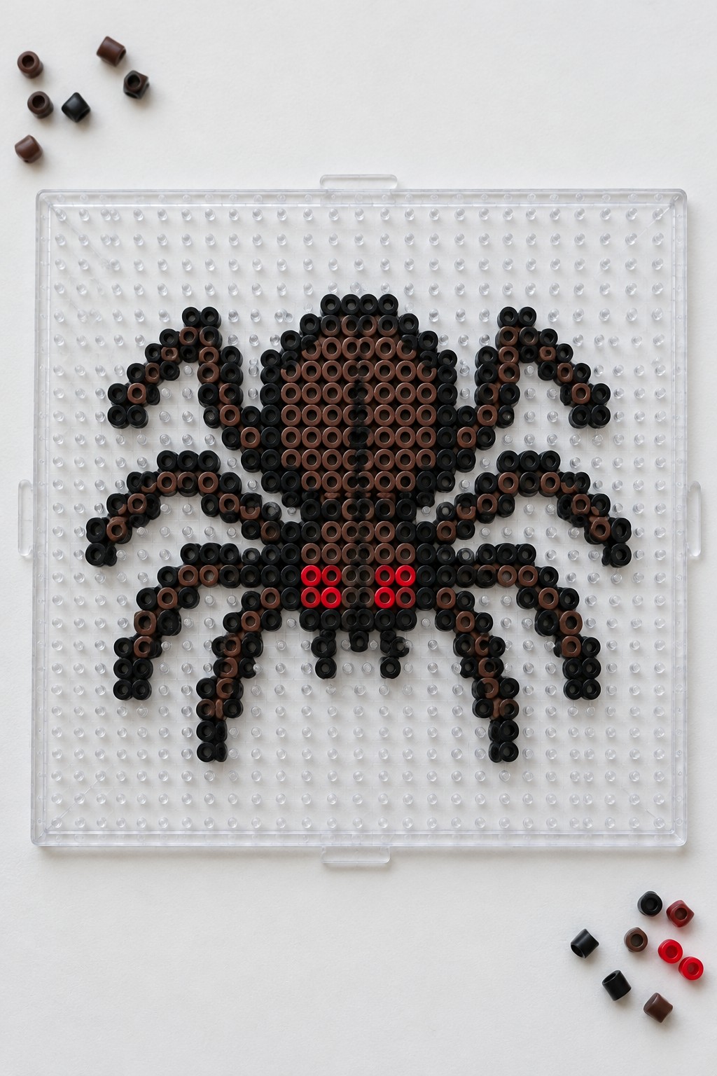 Perler bead spider in black, brown, and red on clear pegboard with loose beads