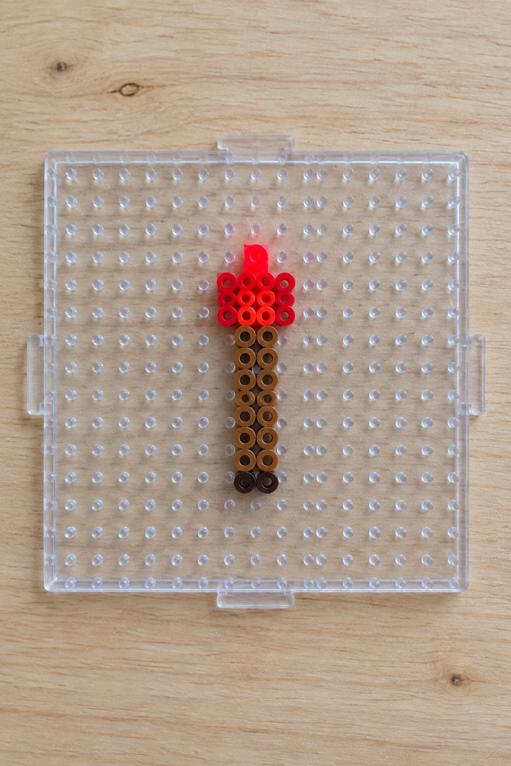 Perler bead arrangement of a Minecraft torch on a pegboard, with red and orange beads forming the flame above a brown shaft.
