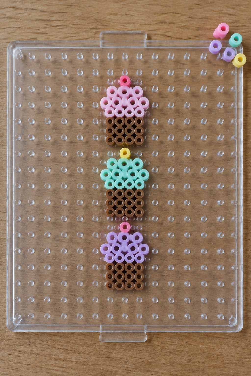 Three perler bead ice cream cones on a clear pegboard with loose beads nearby.