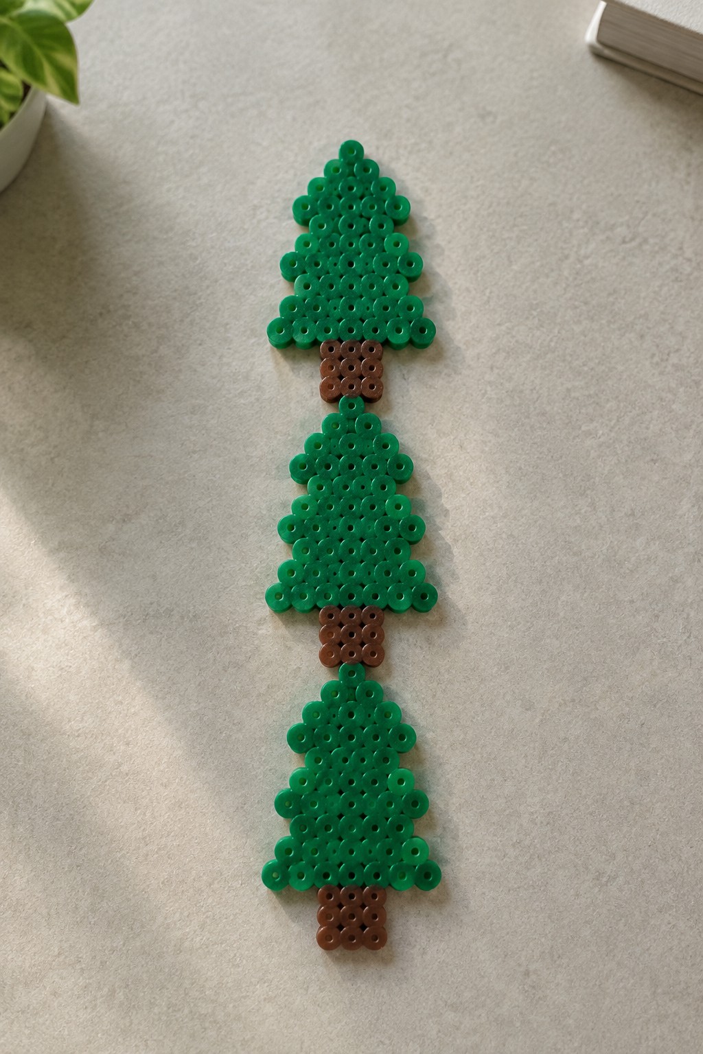 Three green and brown perler bead Christmas trees aligned vertically on a beige surface.