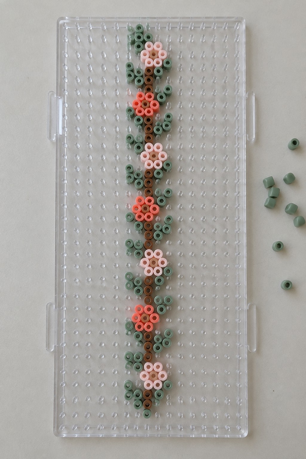 Perler beads form a vertical floral pattern on clear pegboard with loose green beads nearby.