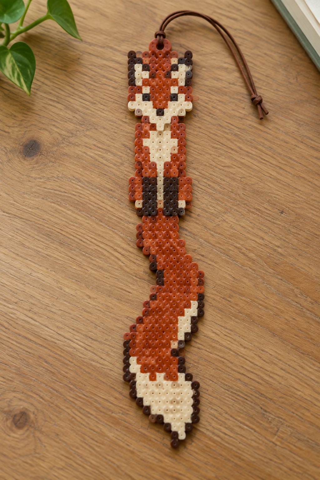 A narrow Perler bead bookmark shaped like a fox using orange, brown, cream, and black beads in a pixel pattern, with a brown cord at the top.