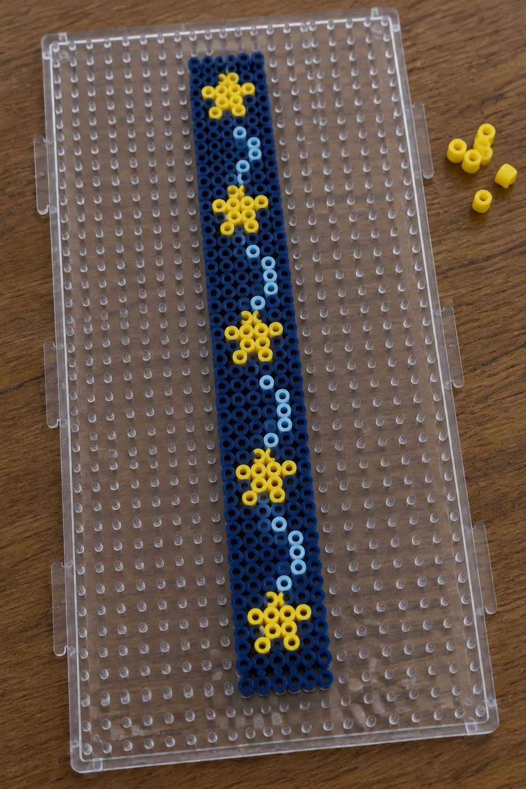 Narrow vertical Perler bead bookmark pattern on a pegboard with dark blue background, yellow stars, and light blue center line.
