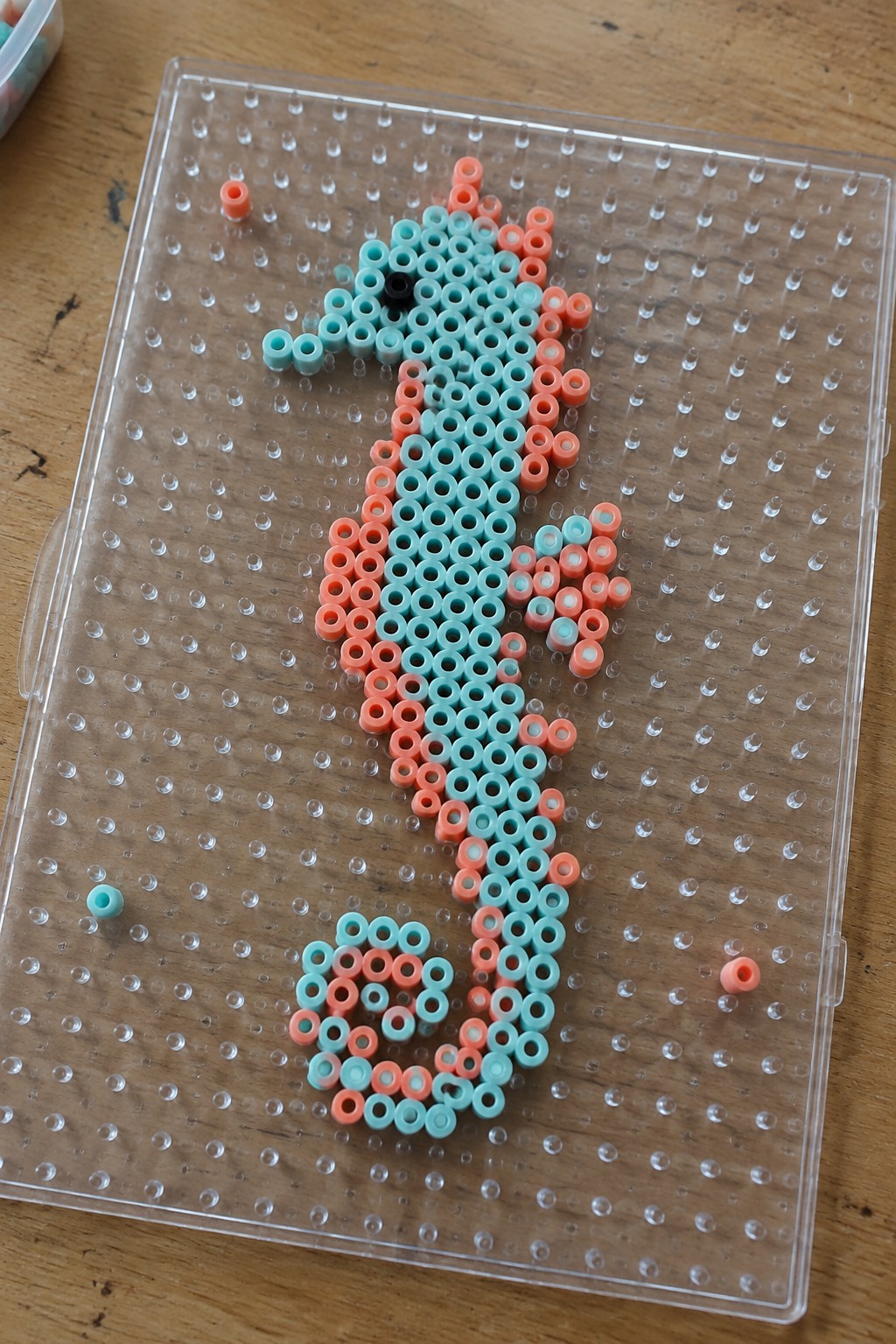 Turquoise and coral perler beads forming seahorse on clear pegboard with loose beads