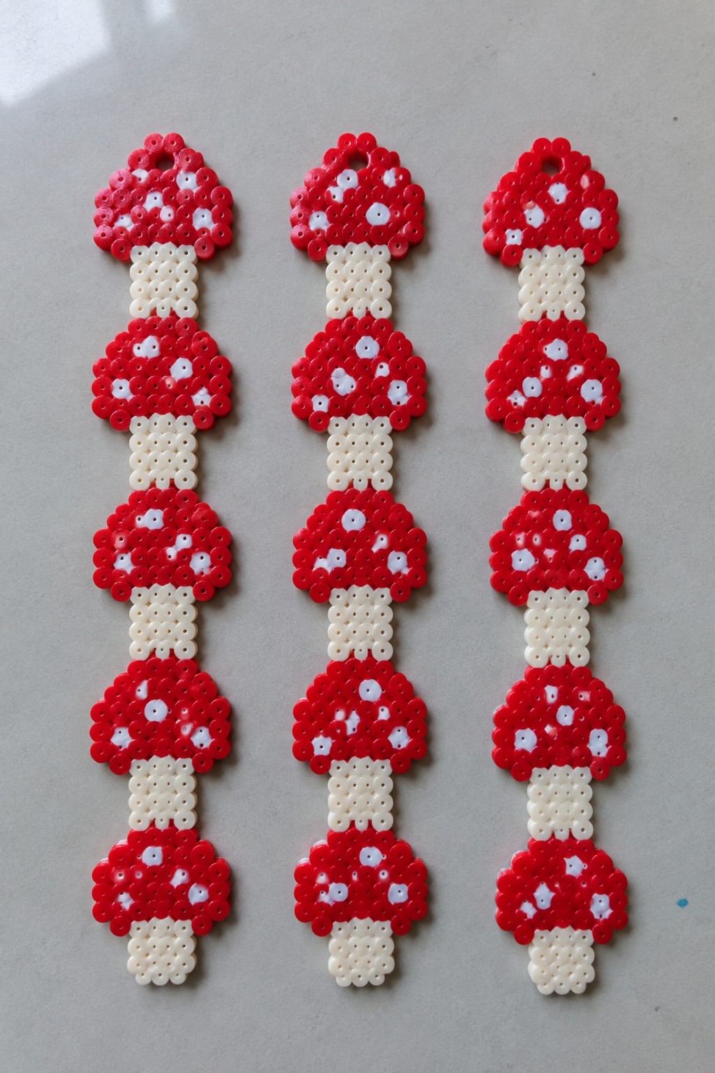 Three vertical red-and-white mushroom perler bead crafts on gray surface.