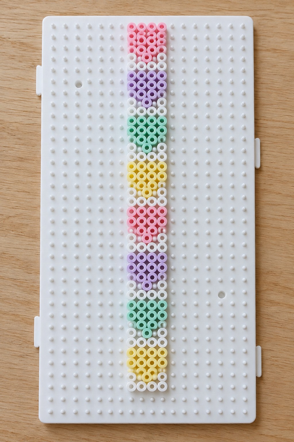 Narrow vertical strip of unfused Perler beads in repeating pink, purple, mint green, yellow, and white bands on a pegboard.