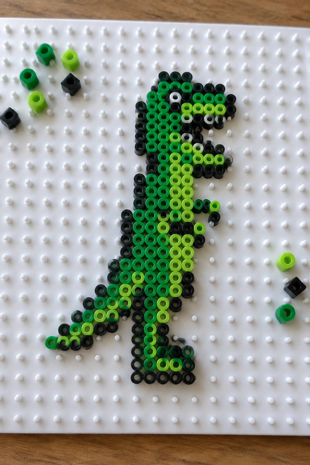 Green and black perler bead dinosaur on white pegboard with loose beads.