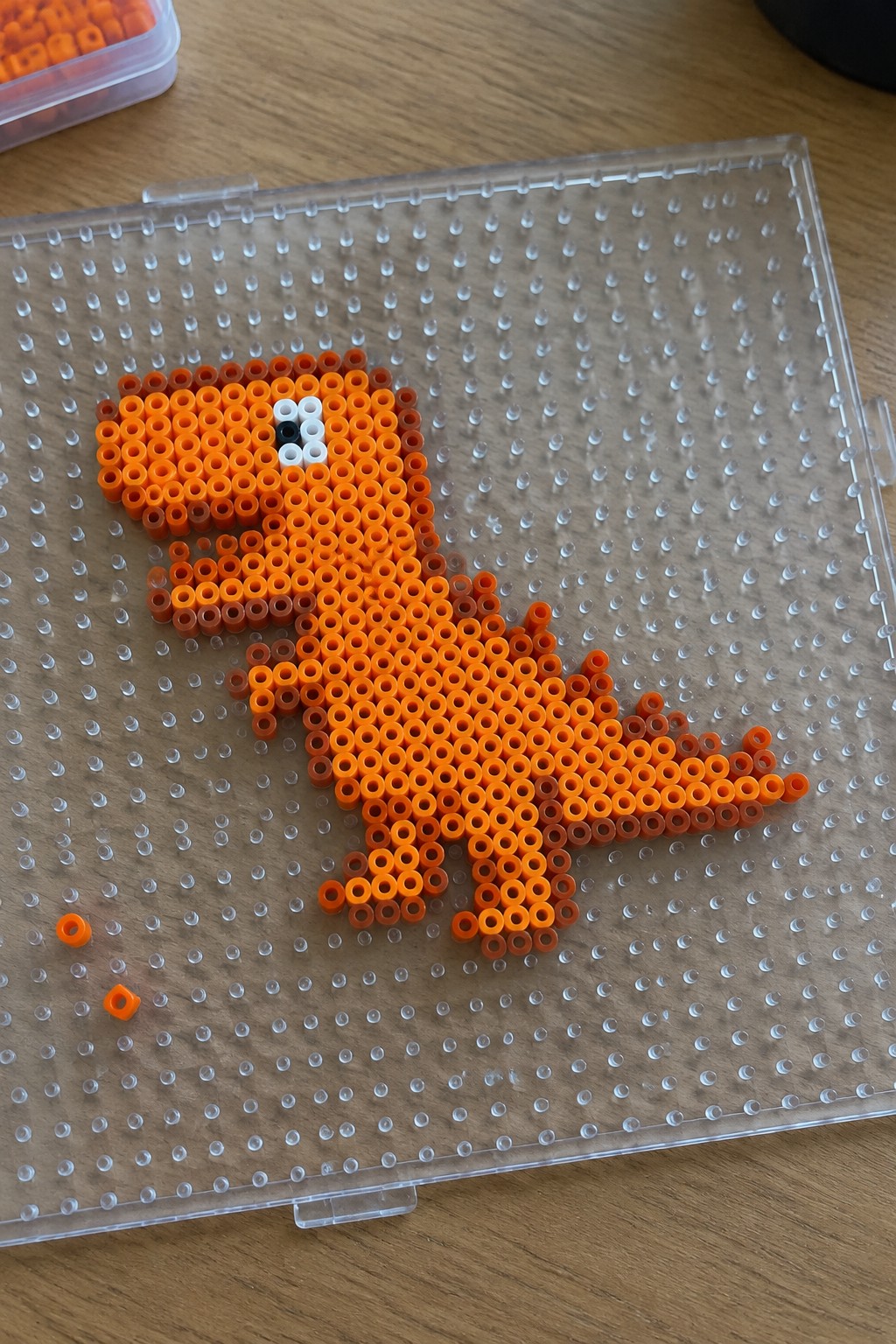 Orange Perler bead T-Rex dinosaur on a clear pegboard with black and white eye.