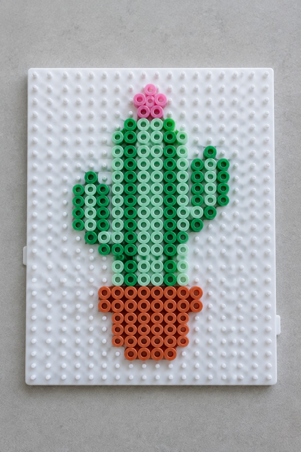 Perler bead cactus with pink flower in an orange pot arranged on a pegboard.
