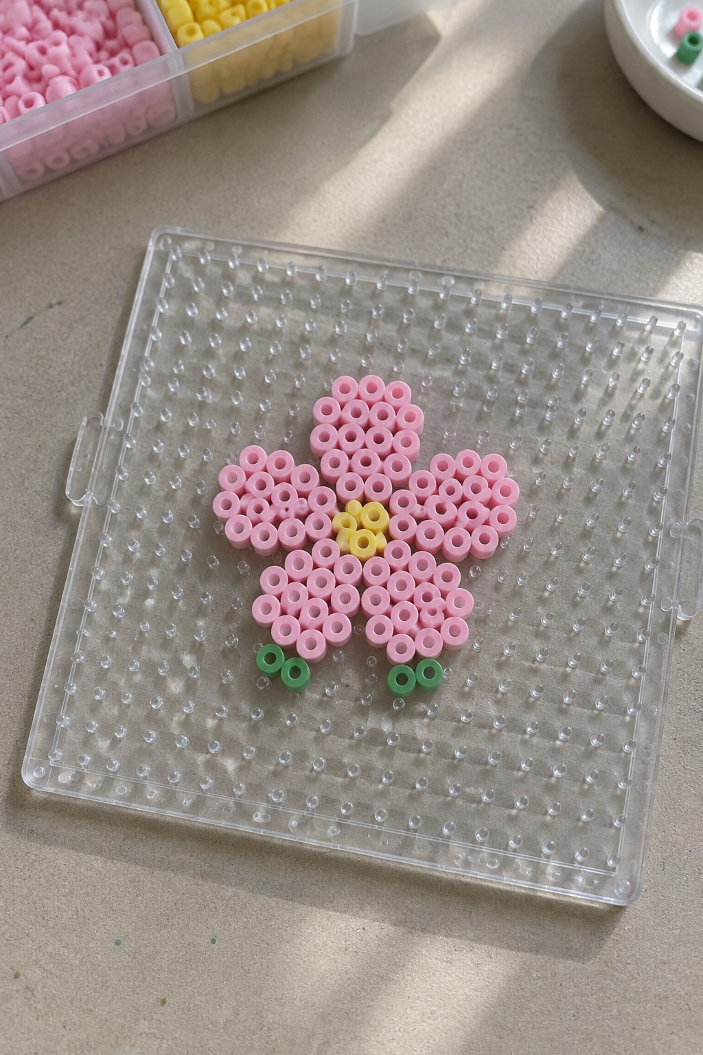 Pink perler bead flower with yellow center on clear pegboard and green accents