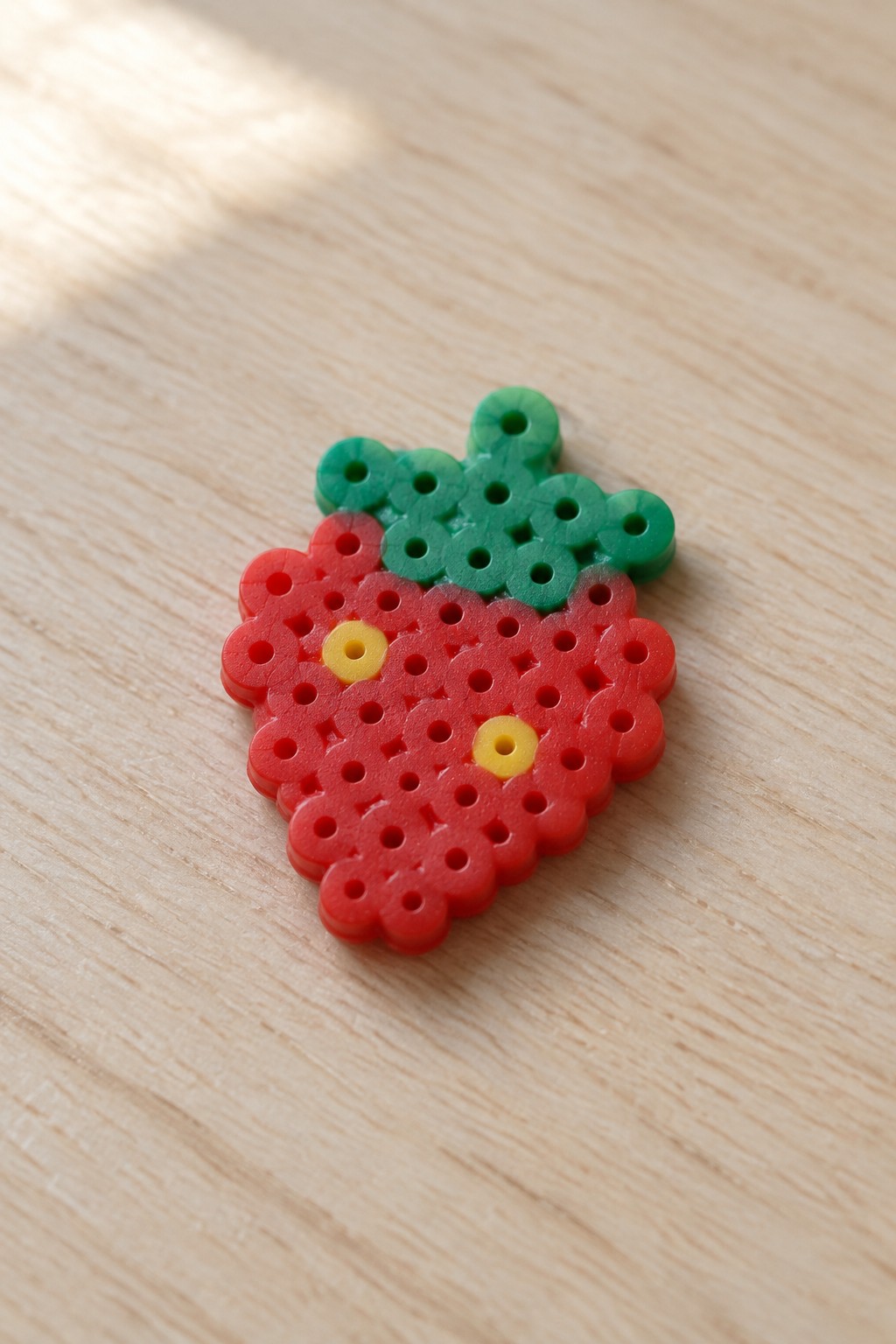 Perler bead strawberry made with red, green, and yellow beads on a wooden surface.