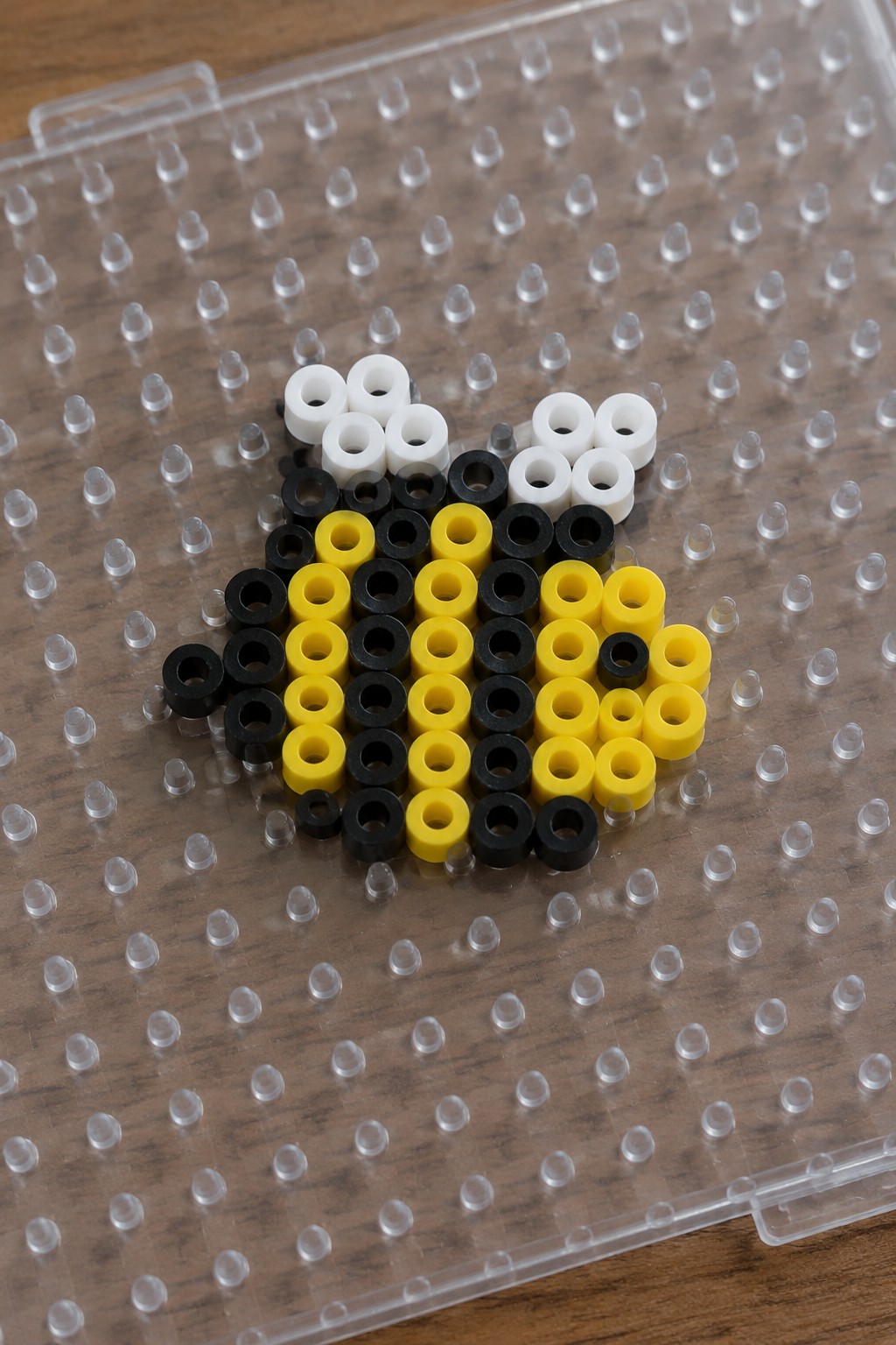 Perler bead bee pattern using black, yellow, and white beads arranged on a clear pegboard.