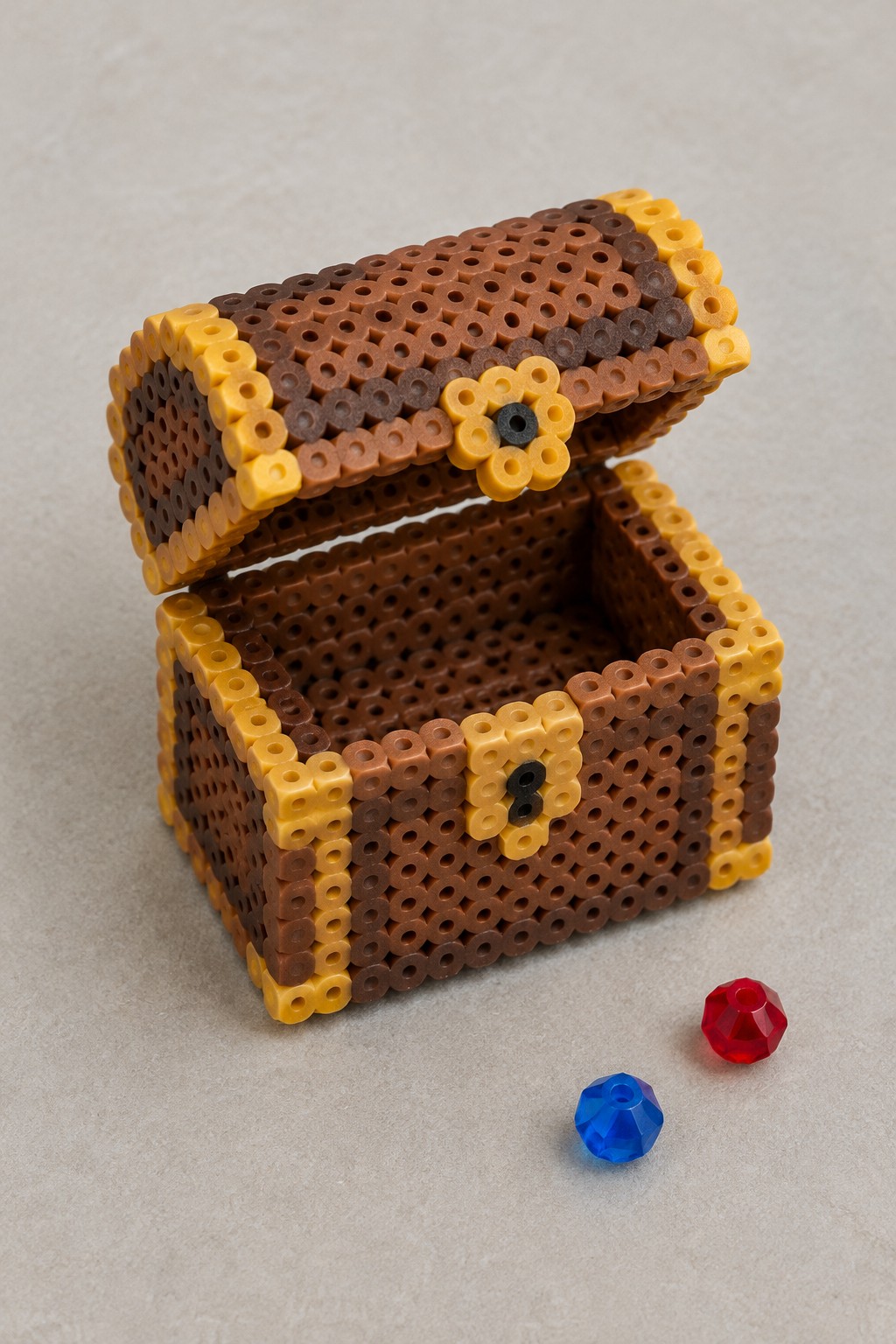 A 3D Perler bead treasure chest in brown and yellow tones with an open lid and two faceted beads next to it.