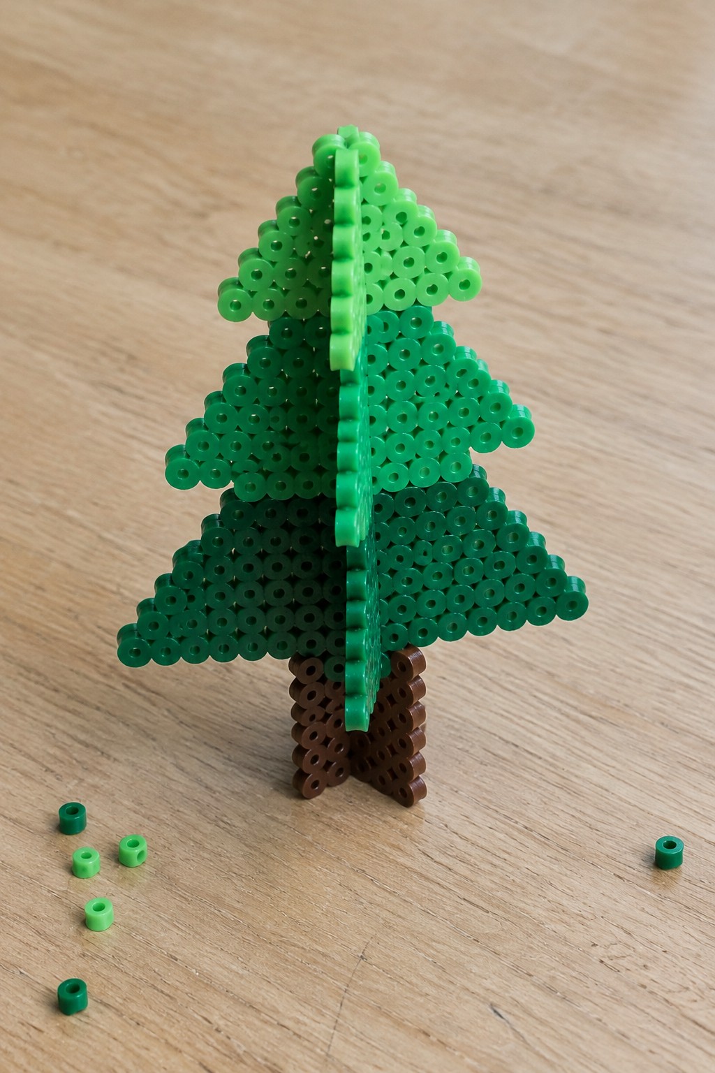 Green perler bead Christmas tree with brown trunk on wooden table amid loose beads