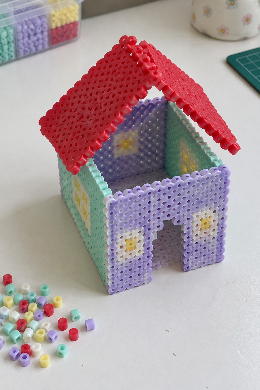 Colorful Perler bead house with red roof, floral windows, and loose beads nearby.