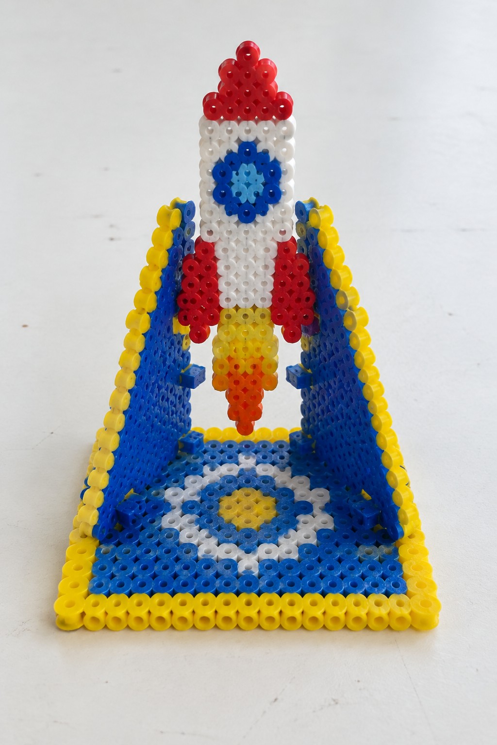 Perler bead rocket with red nose cone lifting off between blue yellow towers on patterned base