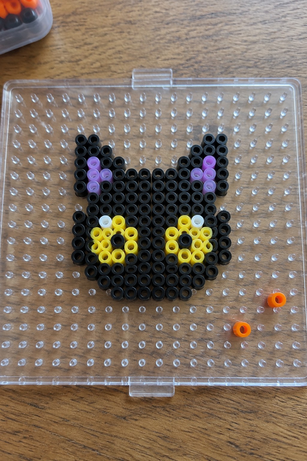 Black cat face made from Perler beads on clear pegboard with purple ears and yellow accents