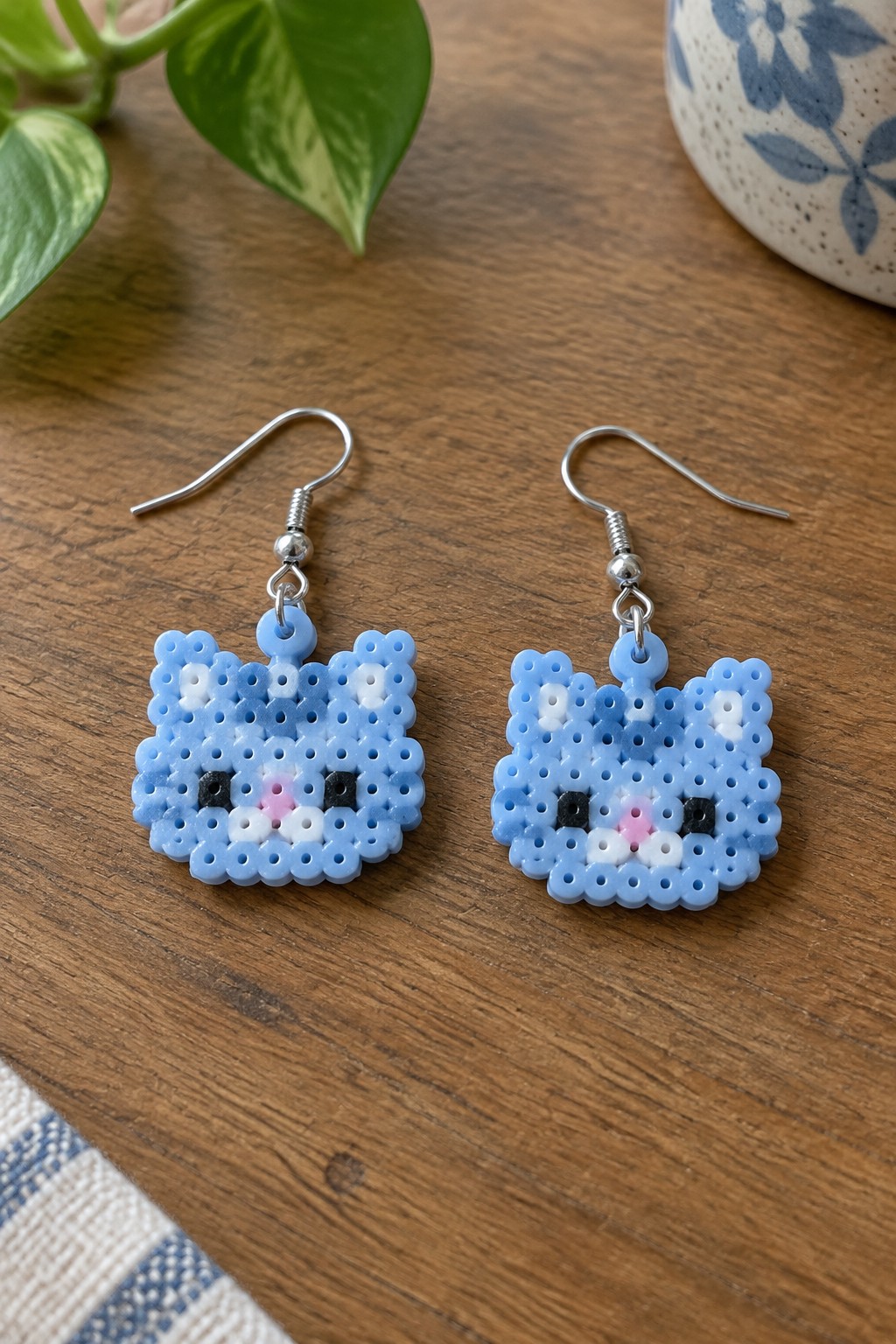 Pair of blue perler bead cat earrings on wooden table with plant