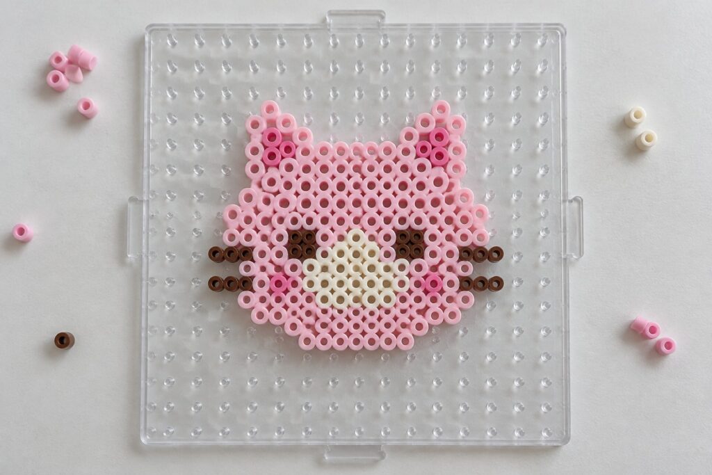 Pink and brown perler beads forming cat face on clear pegboard with loose beads nearby.