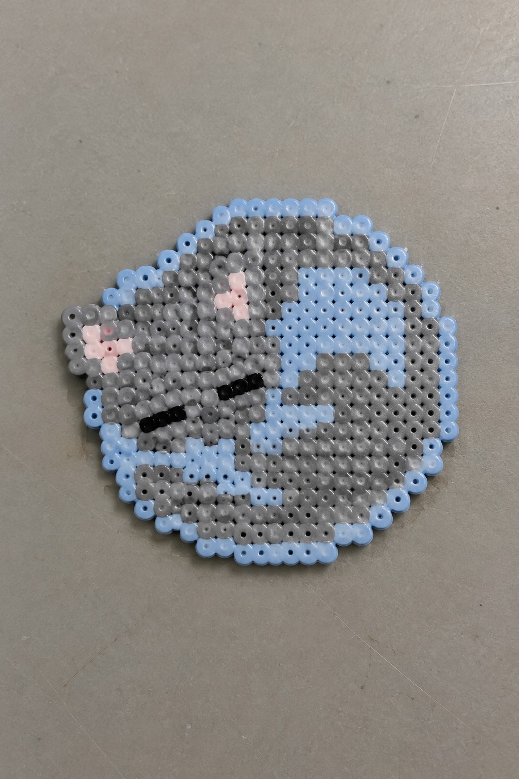 Circular perler bead art of a gray cat face with closed eyes and pink cheeks.