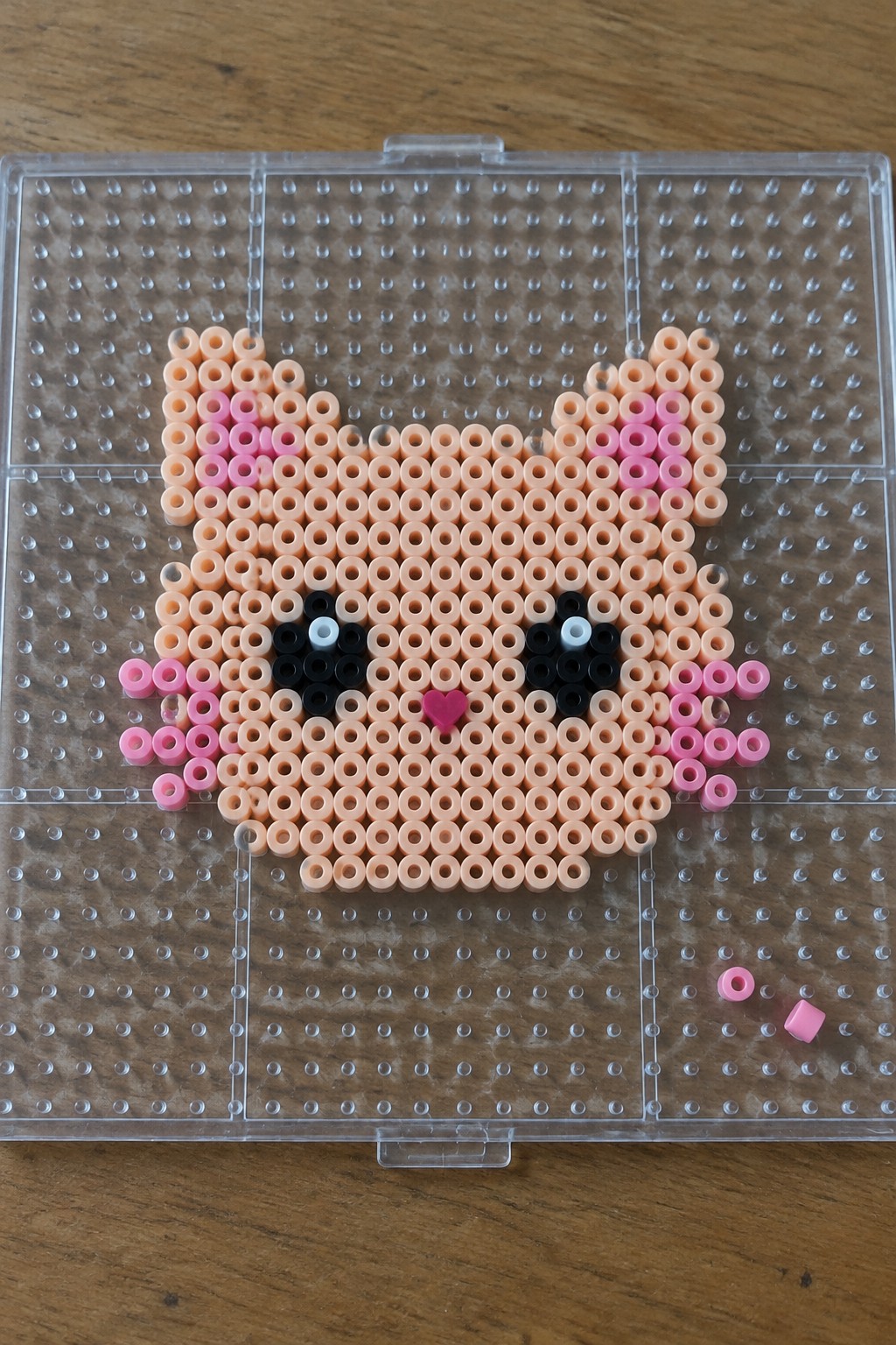 Perler bead cat face with pink cheeks, black eyes, and heart nose on pegboard