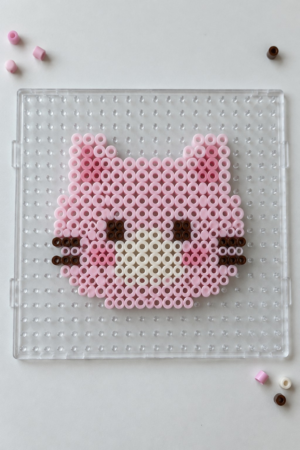 Pink and brown perler beads forming cat face on clear pegboard with loose beads nearby.